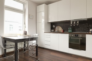 Une cuisine moderne avec des meubles blancs, un tableau noir avec des inscriptions, une bouilloire et une machine à café. Une fenêtre laisse entrer la lumière naturelle et il y a un coin repas avec une table et des chaises.