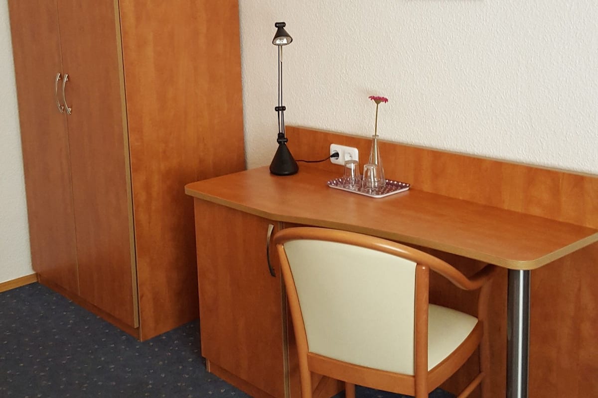 A small room with a wooden desk, a chair, a lamp, and a shelf with a vase and flowers.