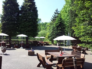Een terras buiten met houten tafels en banken, omringd door bomen.