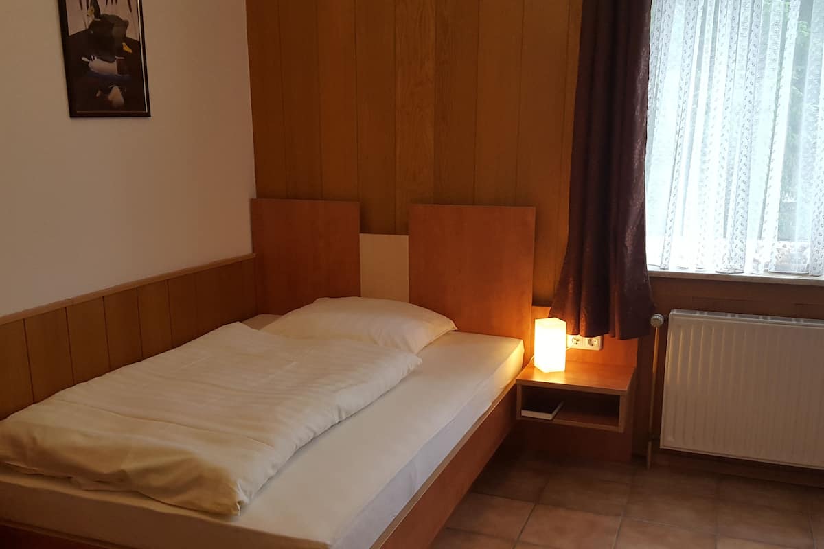 A single bed with a wooden headboard, a bedside table with a lamp, a radiator, and a window with curtains.