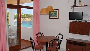 Un coin repas avec une table ronde et des chaises, un téléviseur posé sur un meuble, et une vue sur la mer par une porte vitrée.