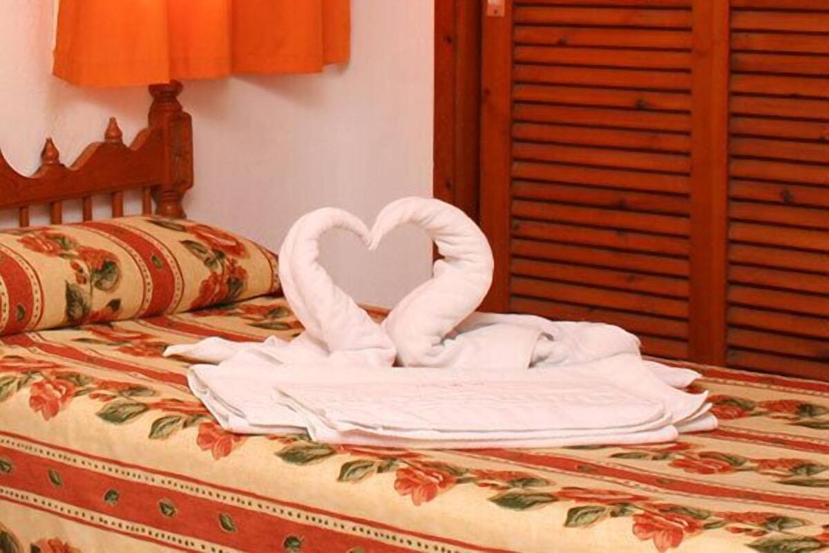A bed with a floral bedspread, a heart-shaped towel arrangement, and an orange curtain.