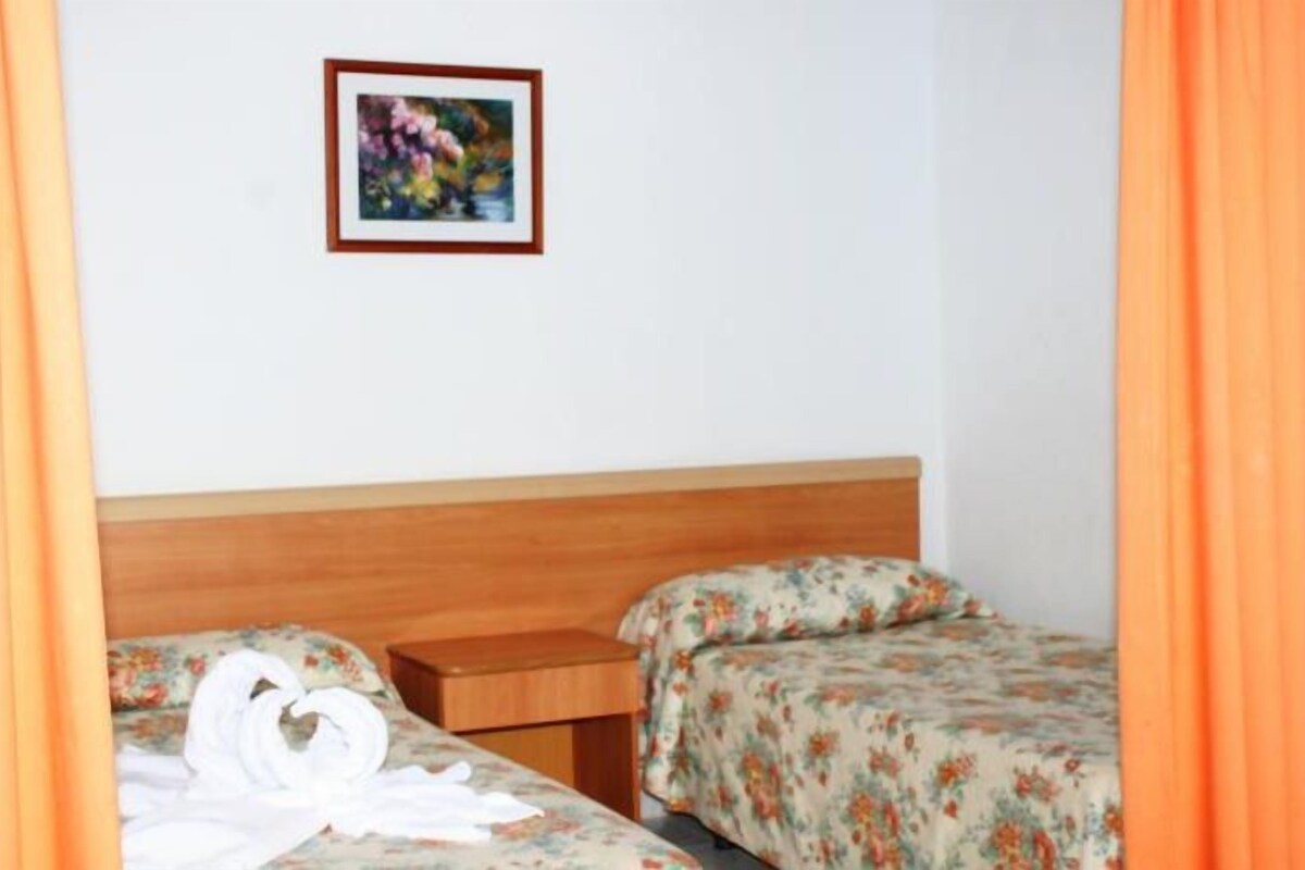 A small, simple room with a wooden headboard, two beds with floral bedding, a small table, and a framed picture on the wall.