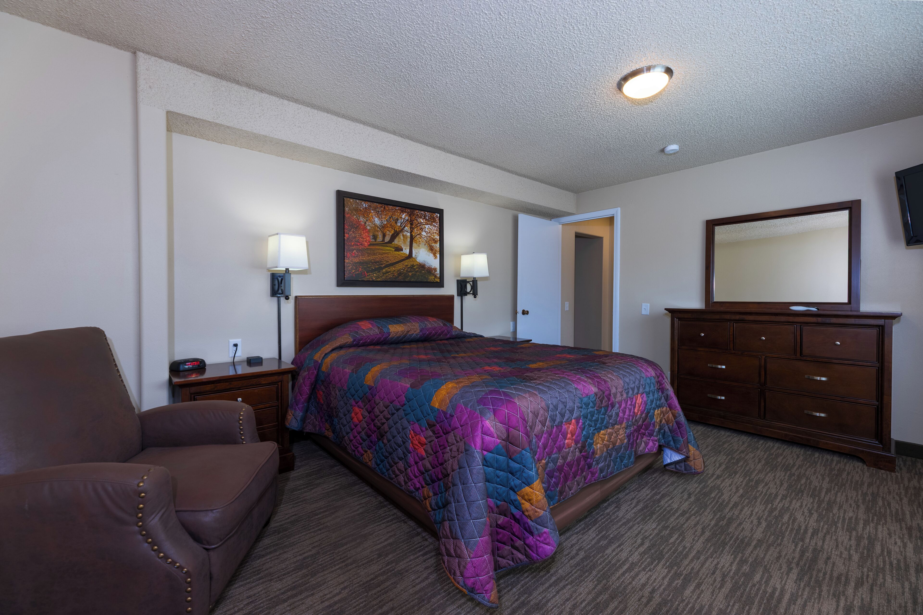 A hotel room with a large bed, a brown leather chair, a dresser, and a painting on the wall.