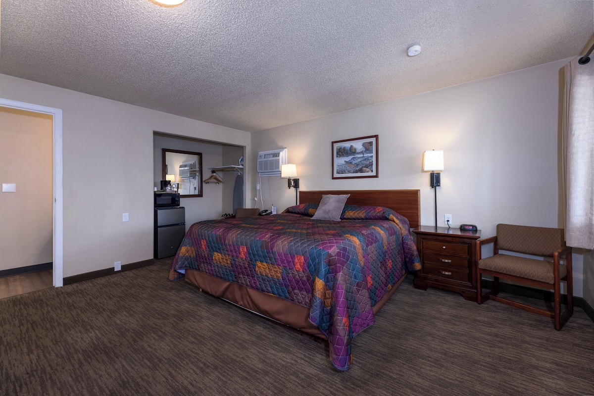 A hotel room with a large bed, a wooden nightstand, a chair, a desk, and a kitchenette in the background.