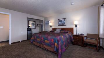 A hotel room with a large bed, a wooden nightstand, a chair, a desk, and a kitchenette in the background.