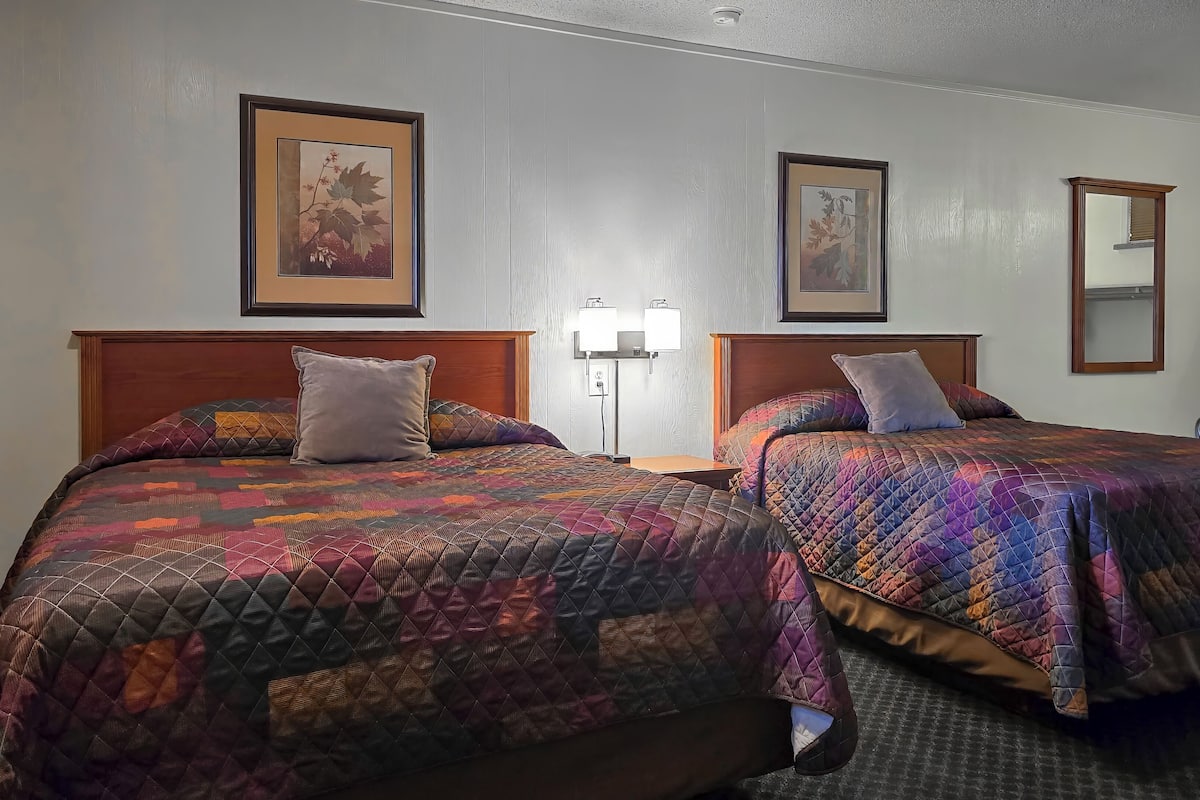 A hotel room with two beds, each with a patterned quilt, a bedside table with a lamp, and framed artwork on the walls.
