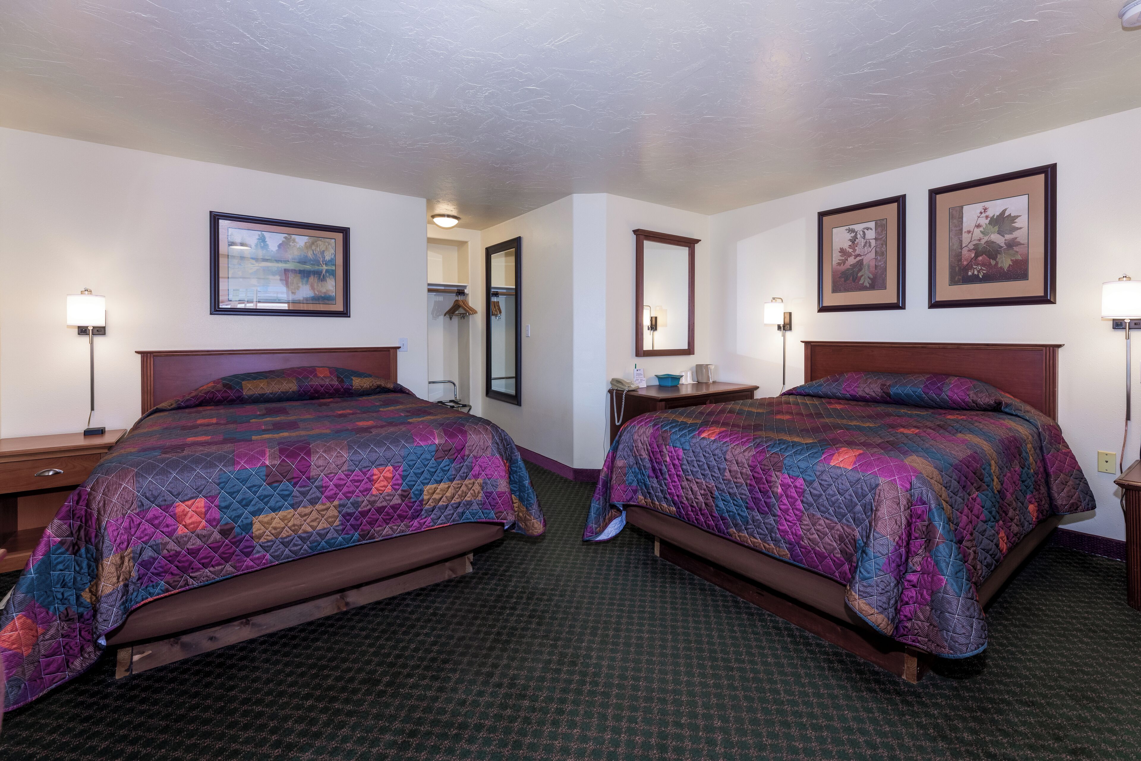 Quarto de hotel com duas camas, cada uma com um colcha colorida, uma mesinha de cabeceira, um espelho e quadros nas paredes.