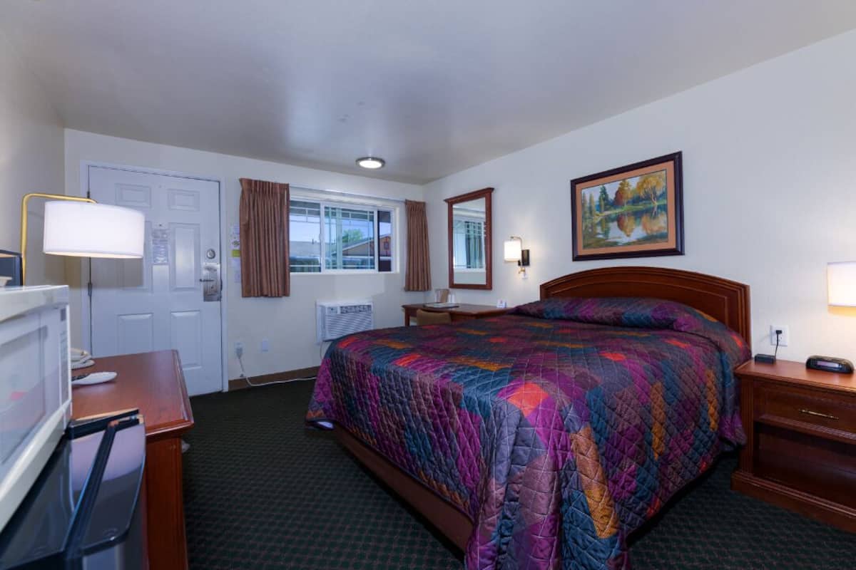 A hotel room with a bed, a desk with a lamp, a television, a window with curtains, and a painting on the wall.
