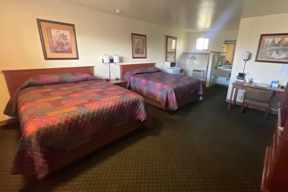 A hotel room with two beds, a desk, a chair, and a small kitchenette.