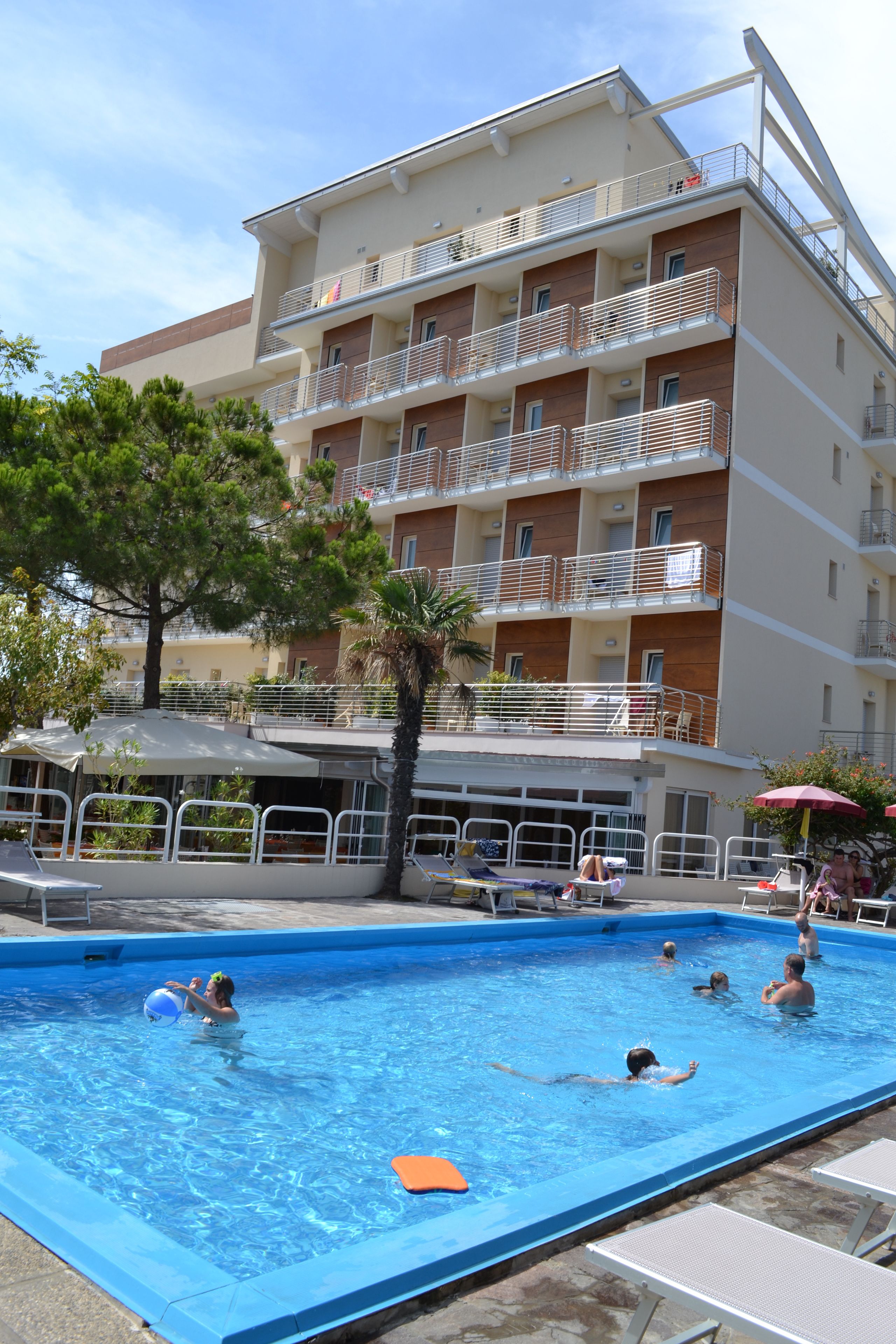 A swimming pool with people swimming and playing, surrounded by a building with multiple balconies and a palm tree.