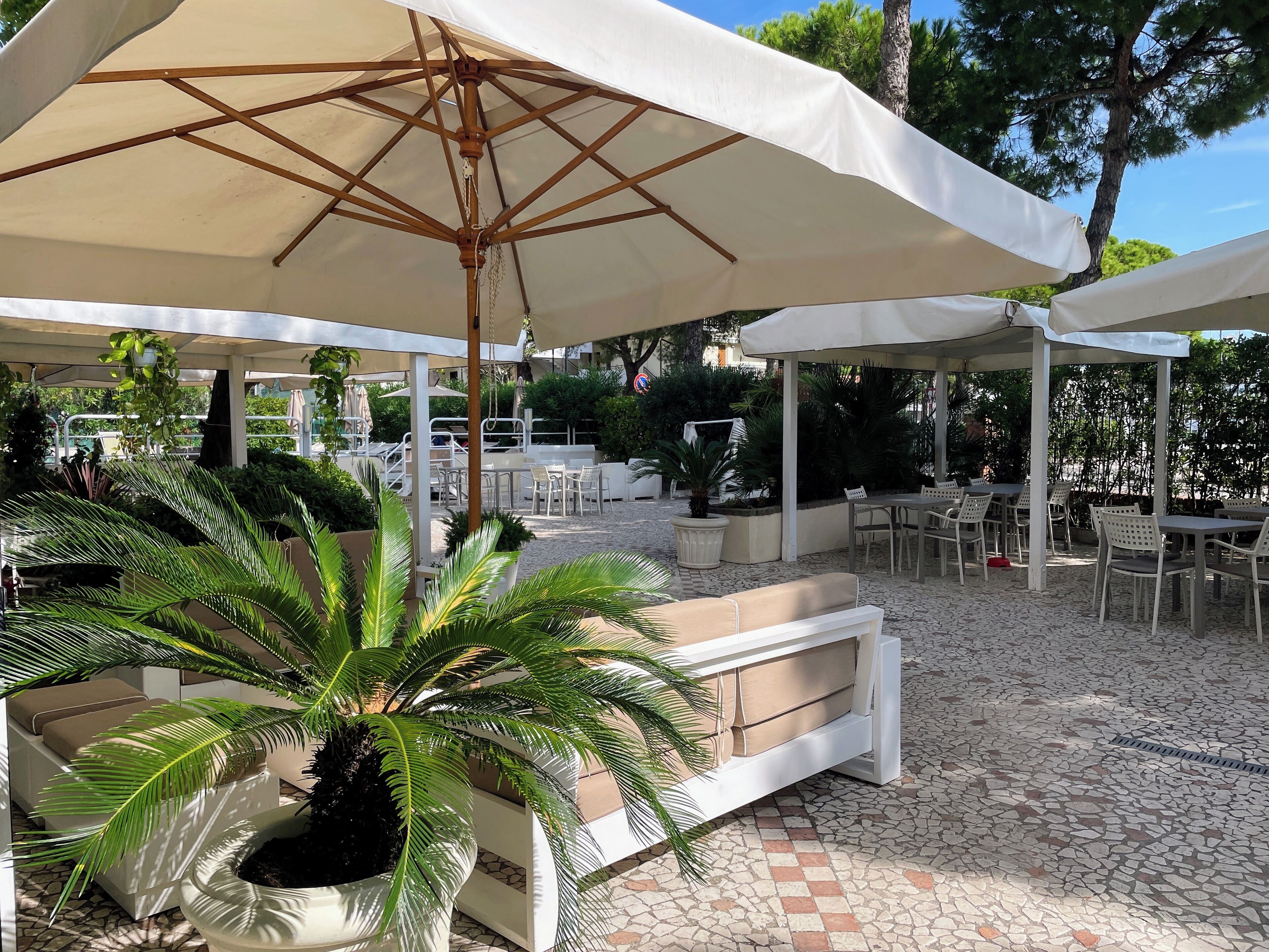 Un espace extérieur aménagé en terrasse, avec des tables et des chaises blanches, protégé par de grands parasols et entouré de plantes et d’arbres en pot.