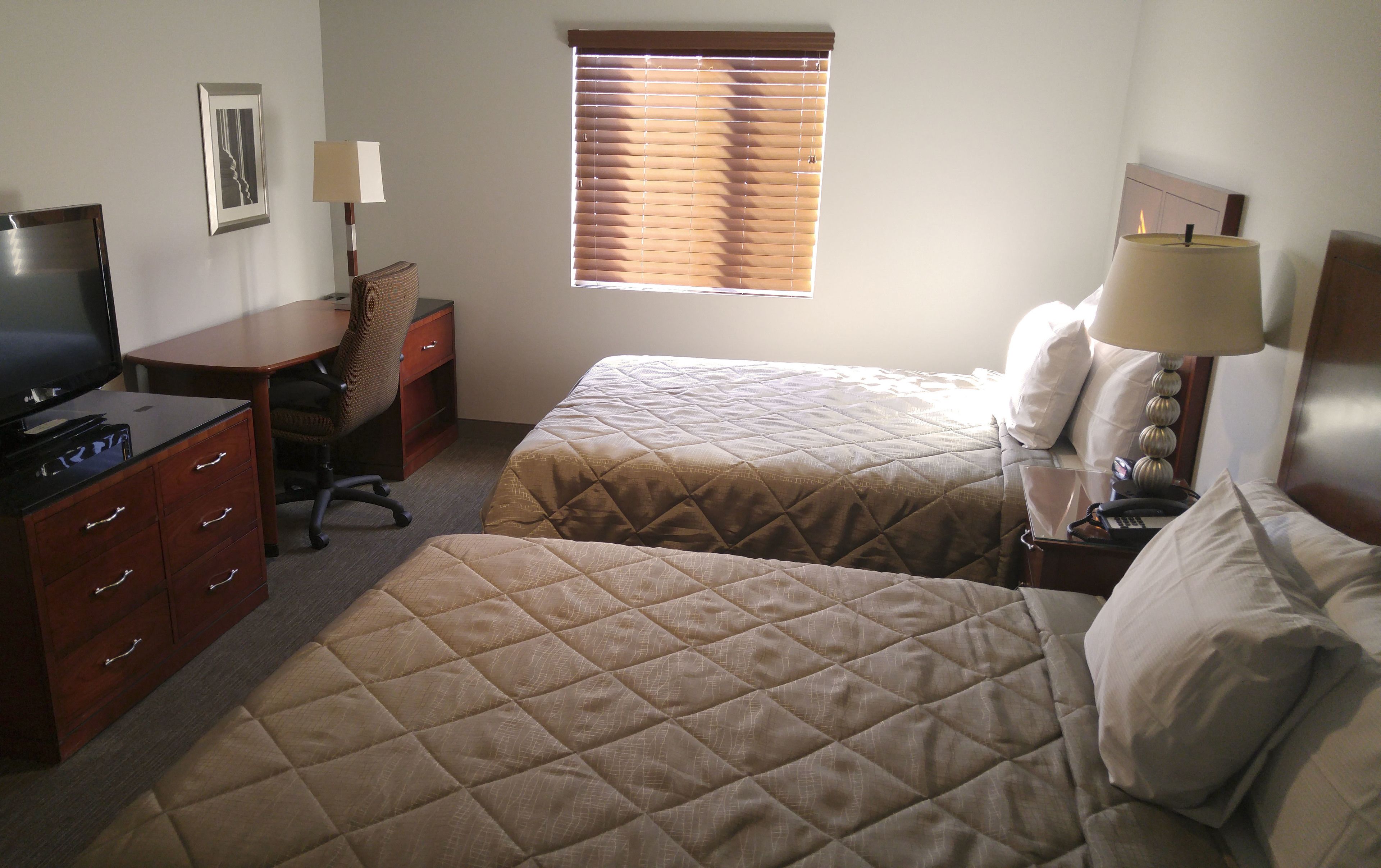 A hotel room with two beds, a desk, a chair, a TV, and a window with blinds.