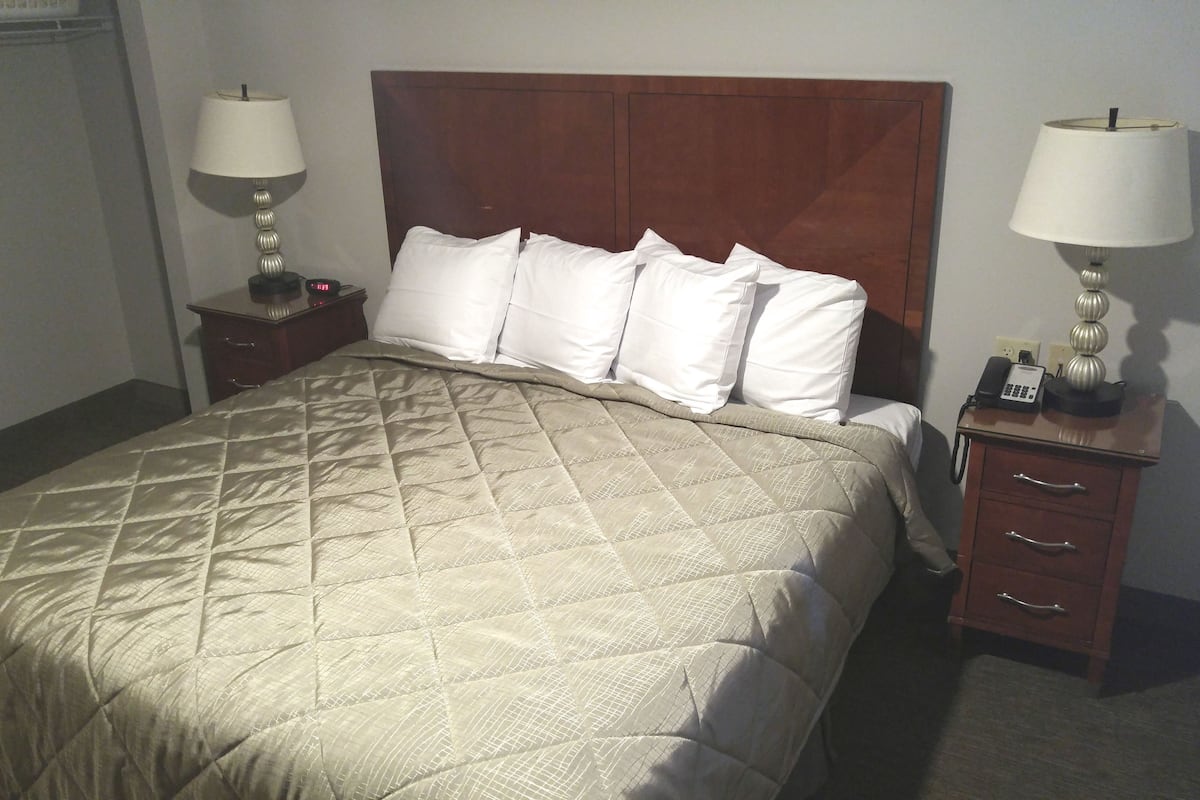 A hotel room with a queen-sized bed, two bedside tables with lamps, a wooden headboard, and a nightstand with a clock.