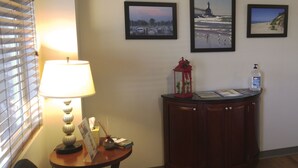 A hotel lobby with a lamp, a small table with a brochure, a cabinet, and three framed pictures on the wall.