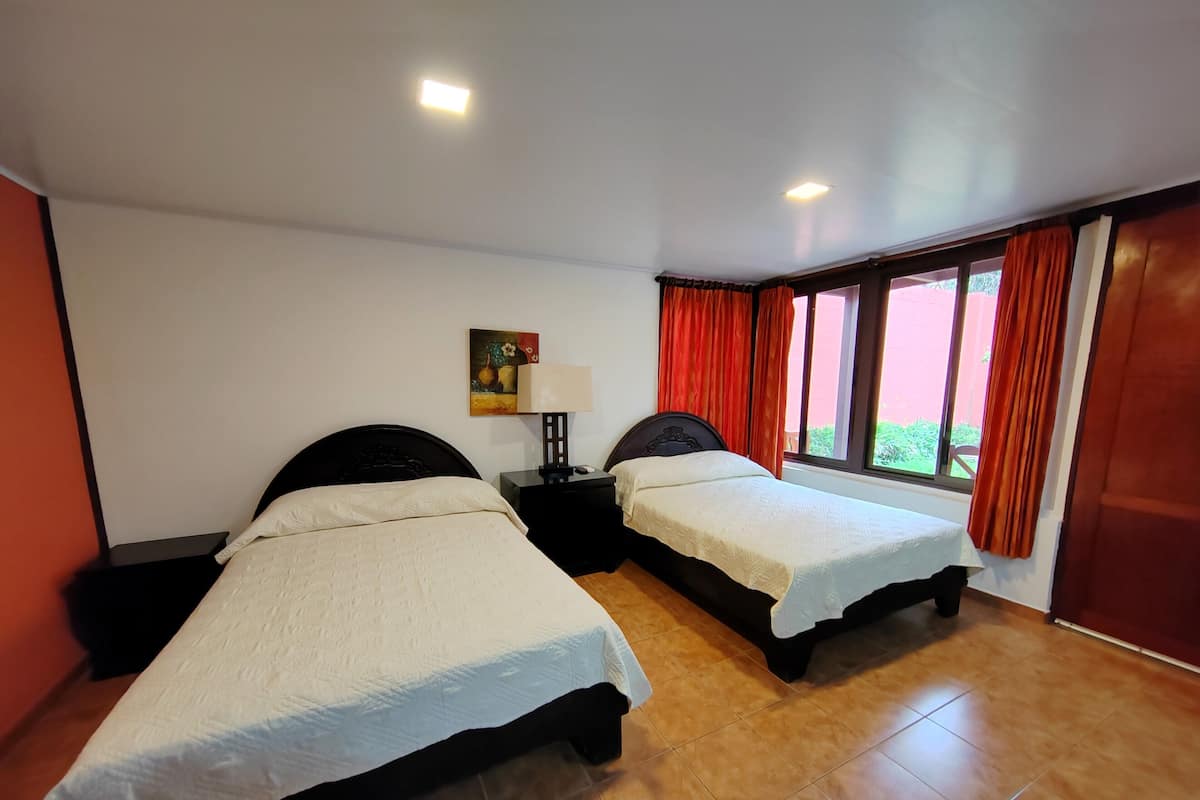 A hotel room with two single beds, a nightstand with a lamp, a painting on the wall, and a window with red curtains.