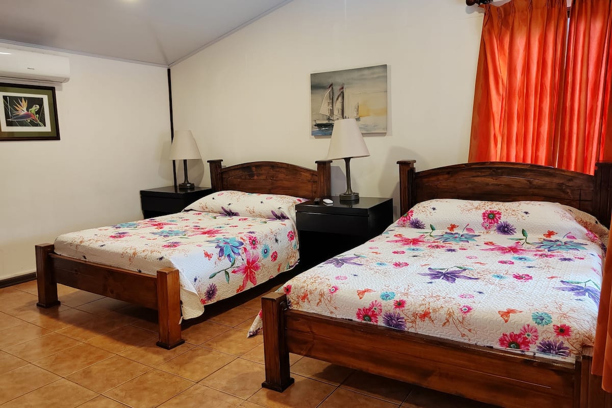 A room with two wooden beds, each with a floral bedspread, a nightstand with a lamp, a painting on the wall, and orange curtains.