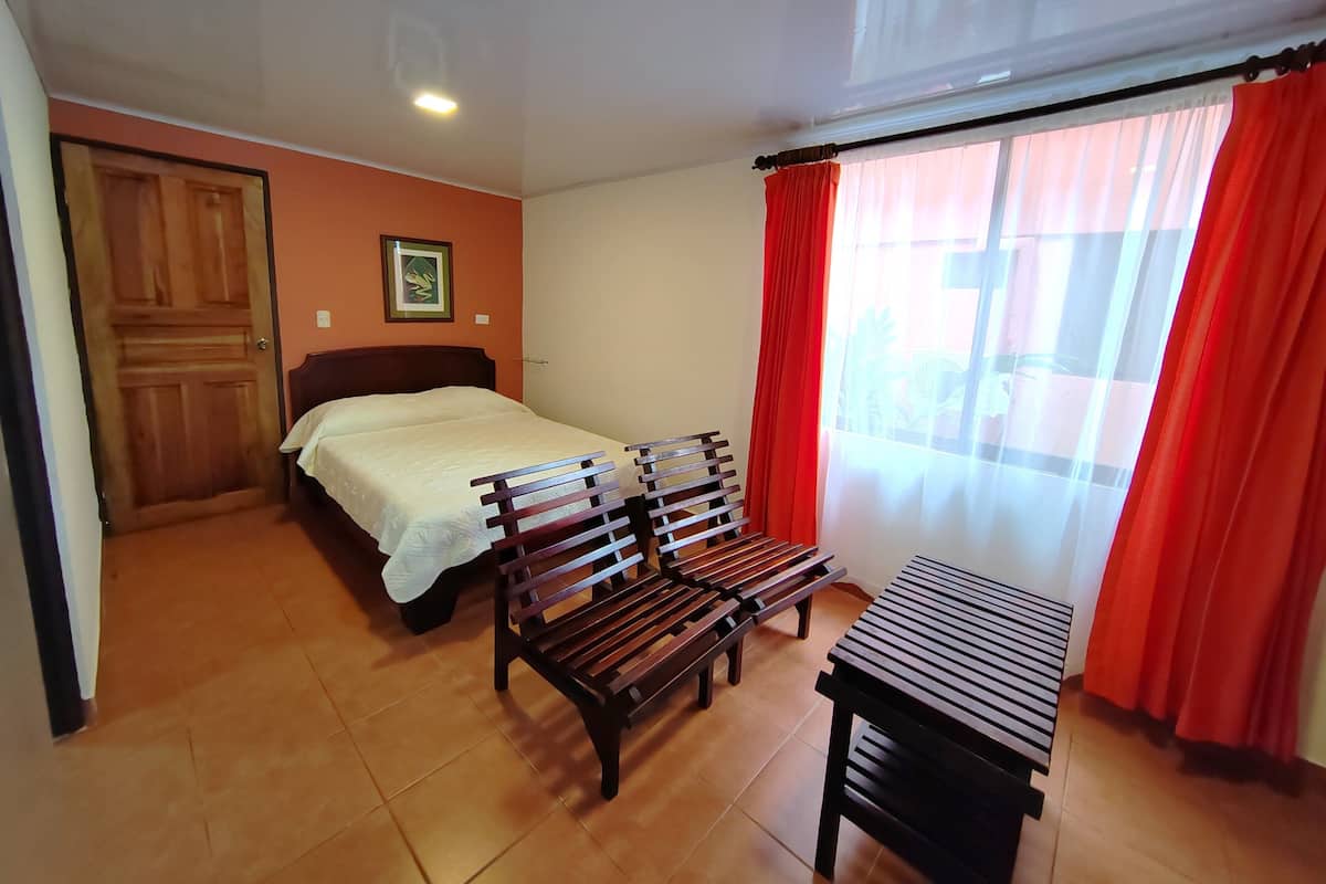 A bedroom with a bed, a wooden bench, a small table, a door, and a window with red curtains.