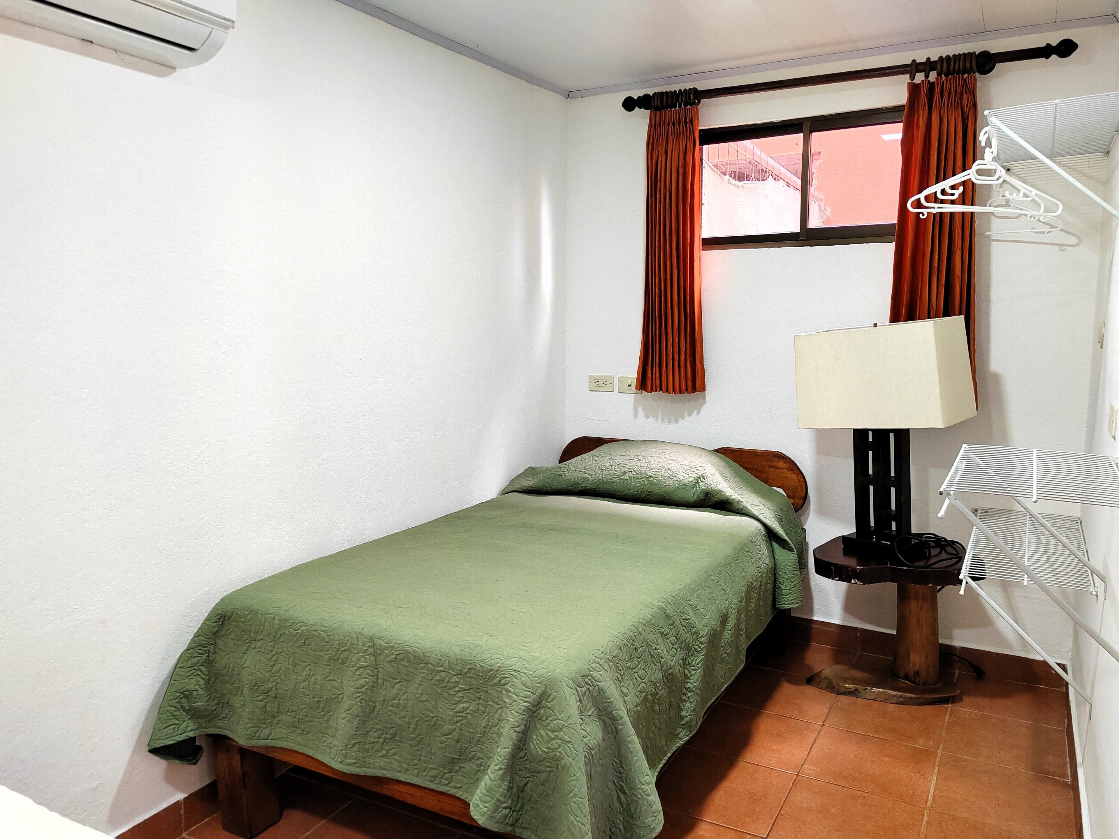 A small, neatly arranged bedroom with a single bed, a nightstand, a lamp, and a window with curtains.