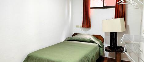 A small, neatly arranged bedroom with a single bed, a nightstand, a lamp, and a window with curtains.