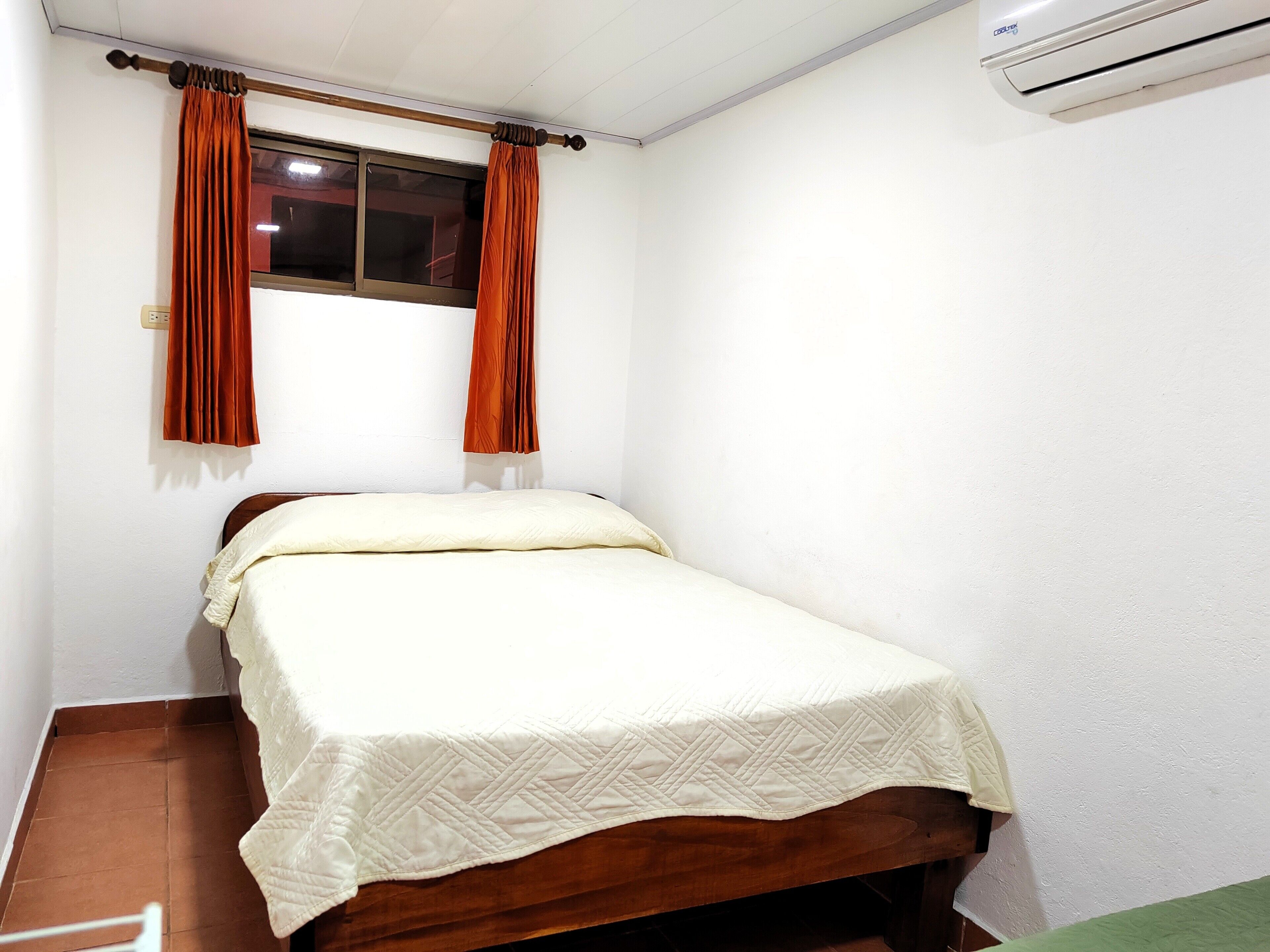 Una habitación pequeña con una cama individual, cabecera de madera, ventana con cortinas marrones y un equipo de aire acondicionado en la pared.