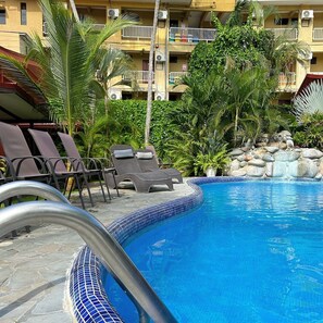 A swimming pool with lounge chairs and a stone deck, surrounded by tropical plants and a multi-story building with balconies. - Hotel Los Ranchos (Jacó)