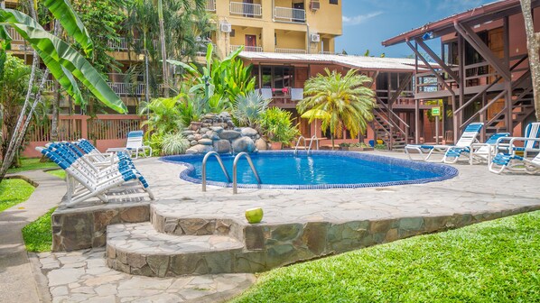 A swimming pool with lounge chairs, a stone pathway, and a building with balconies in the background.