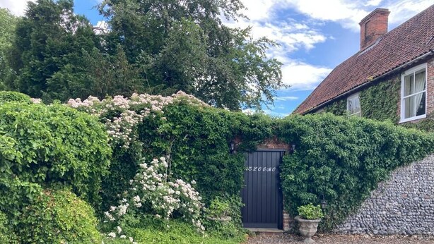 A traditional house with a dark wooden door, surrounded by lush greenery and flowering plants. - Grange Farm House (Thetford)