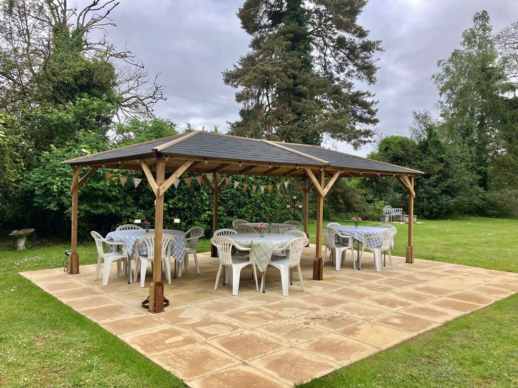 A covered outdoor seating area with a pergola, white plastic chairs, and a stone patio.