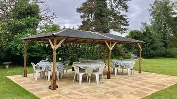A covered outdoor seating area with a pergola, white plastic chairs, and a stone patio.