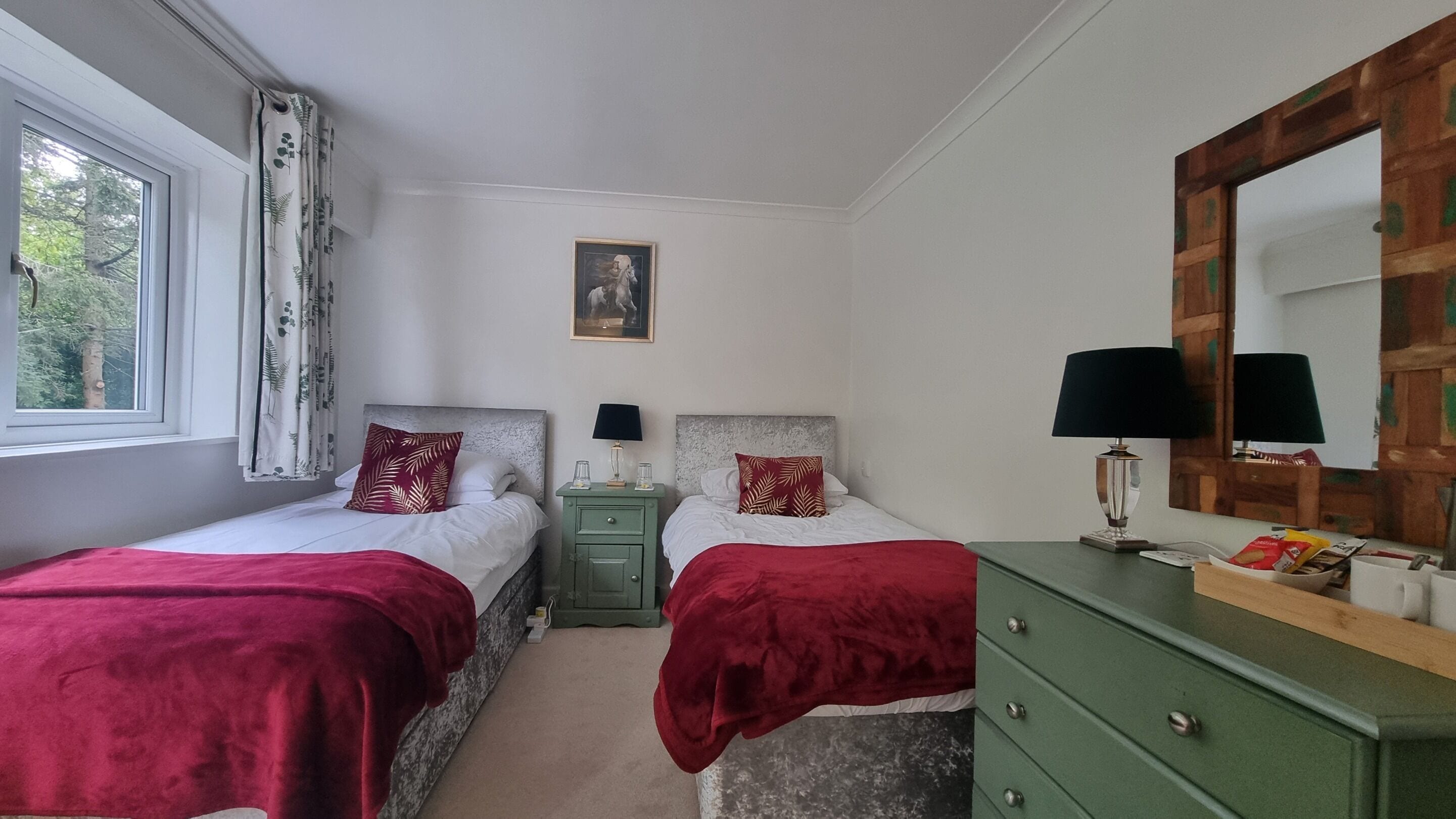 A bedroom with two beds, each with a red blanket, a green nightstand with a lamp, and a wooden dresser with a mirror.