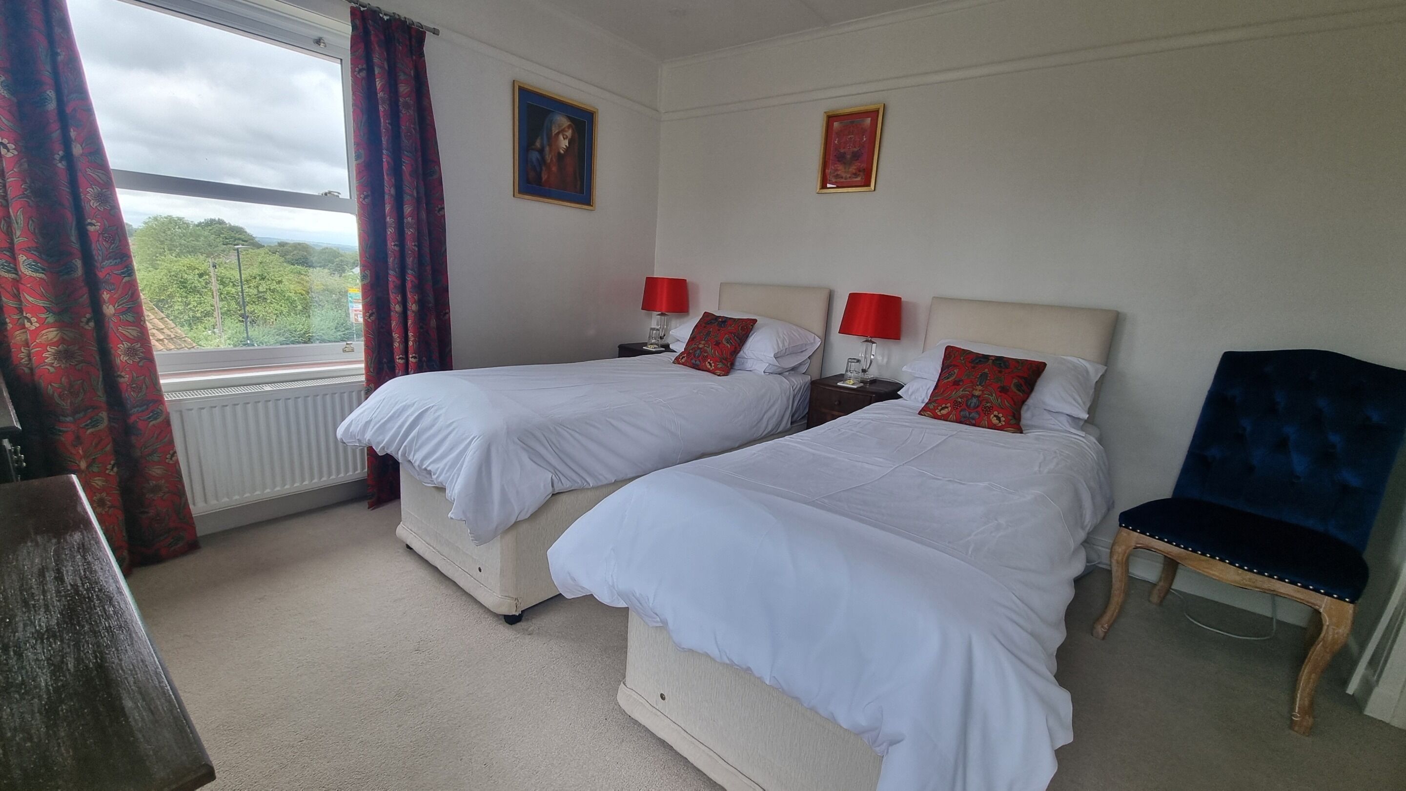 A hotel room with two beds, red lamps, a blue chair, and a view of greenery outside.