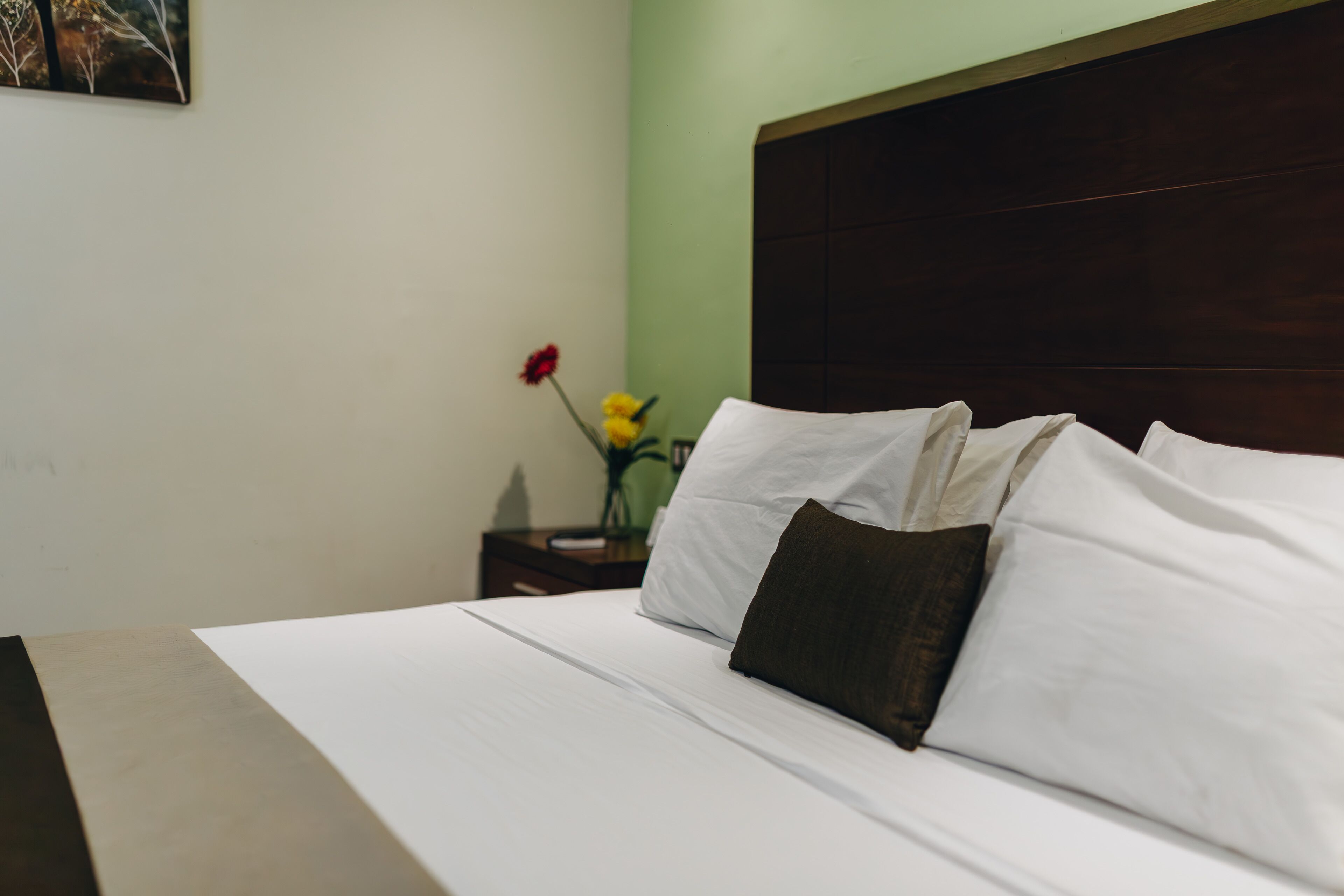 A neatly made bed with white linens and a dark brown pillow, a wooden headboard, and a small table with a vase of flowers.