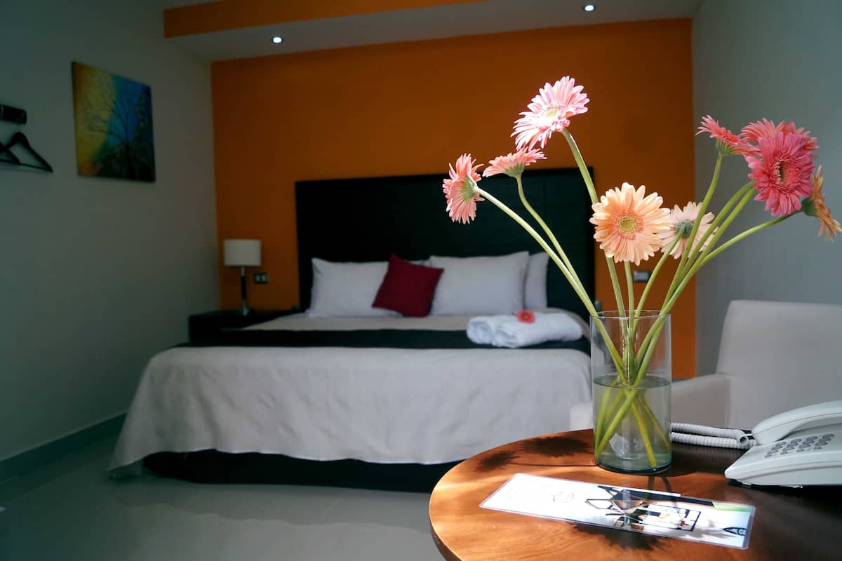 Habitación de hotel con cama, mesita de noche con teléfono y folleto, y un florero con flores.