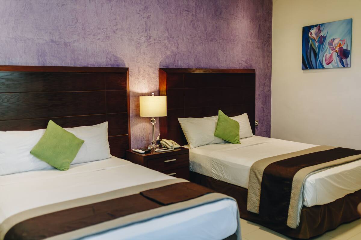 Habitación de hotel con dos camas, cada una con un almohadón y una mesita de noche con lámpara.