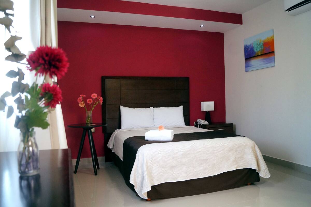 Una habitación de hotel con una cama grande, una mesita de noche, un florero con flores y un cuadro en la pared.