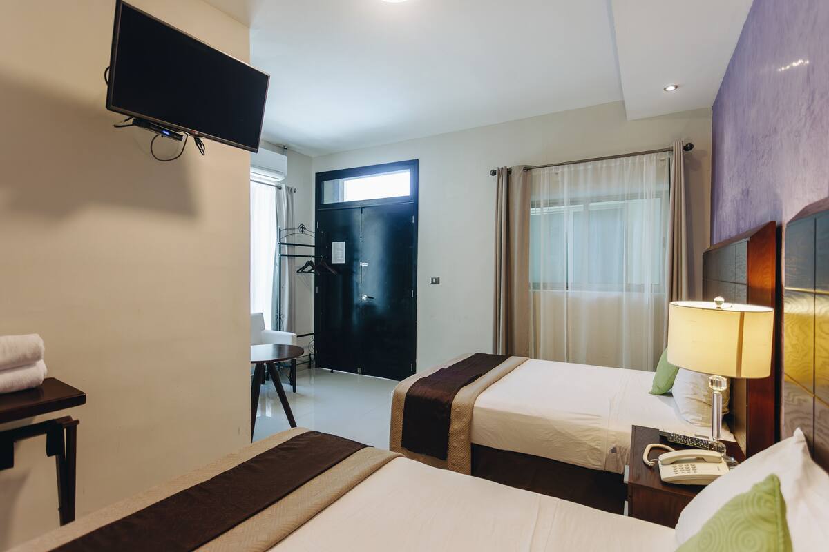 Habitación de hotel con dos camas, un televisor montado en la pared, un escritorio con una silla, una puerta que da al baño y una ventana con cortinas.