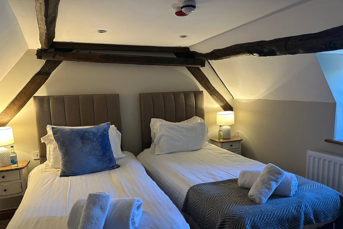 A bedroom with two beds, wooden beams, a nightstand with a lamp, and a window.