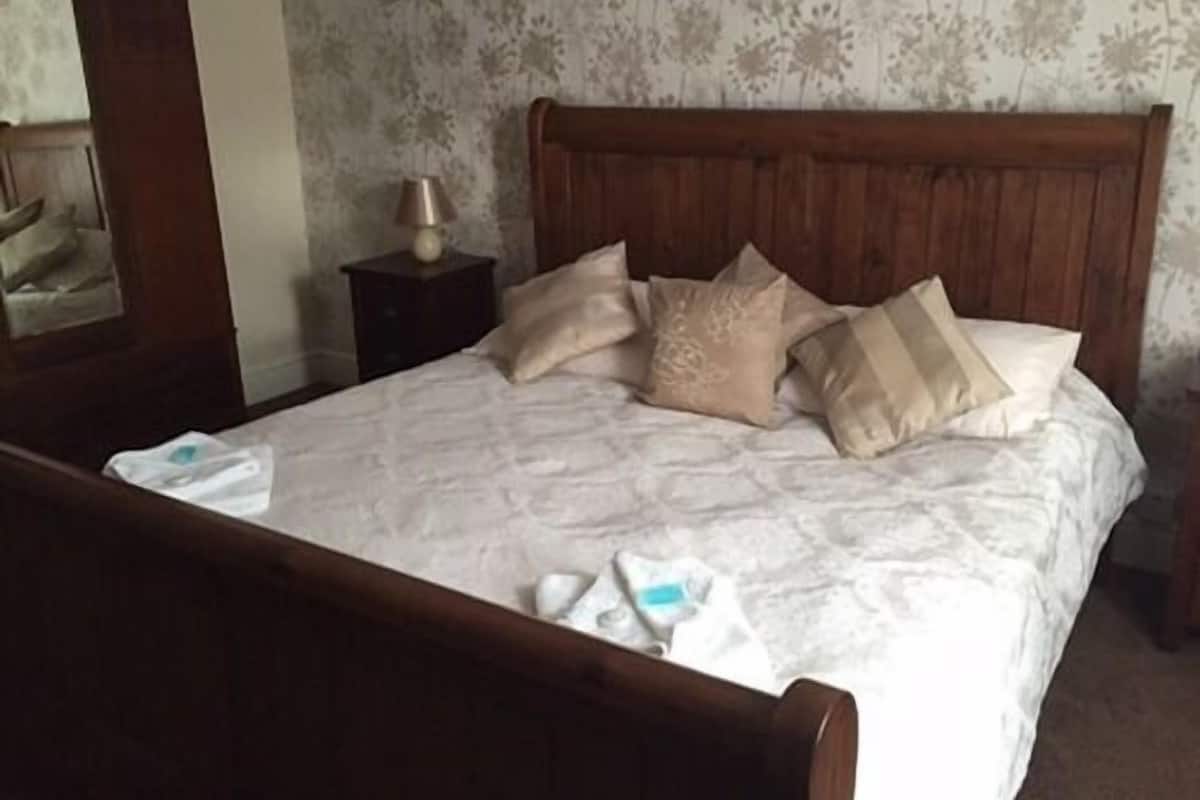 A neatly made bed with a wooden headboard, a bedside table with a lamp, and a floral wallpaper pattern.
