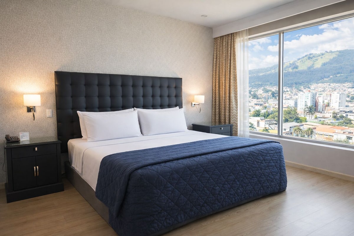 A hotel room with a large bed, two bedside tables, a city view, and a window with curtains.