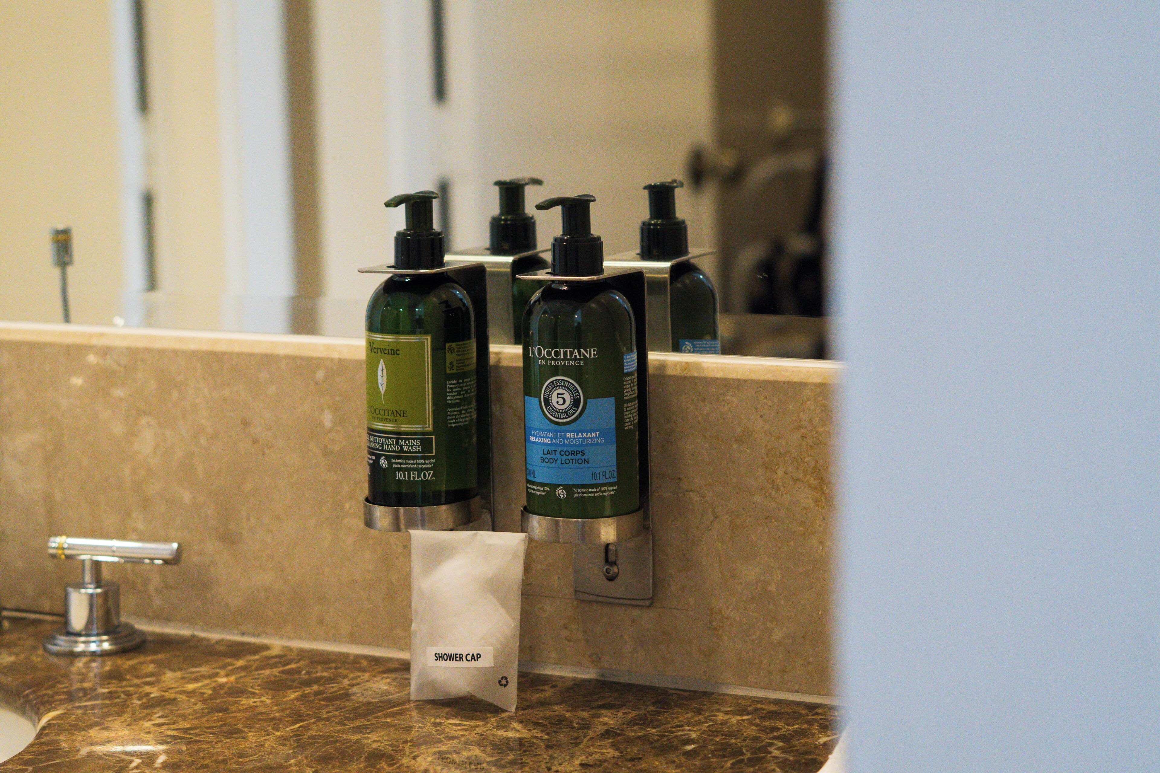 Een badkamer met een marmeren aanrechtblad, een dispenser voor douchemutsen en twee flesjes L'OCCITANE body lotion.