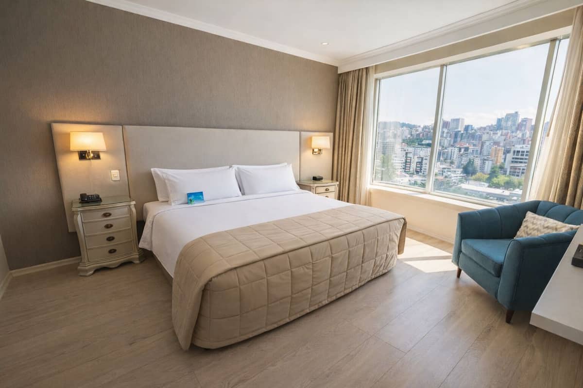 A hotel room with a large bed, two bedside tables, a nightstand, a blue armchair, and a large window with a city view.