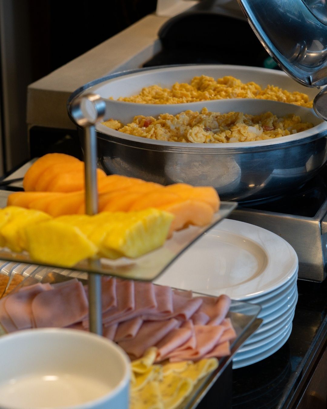 A buffet with scrambled eggs, orange slices, pineapple chunks, ham, and a bowl of soup.