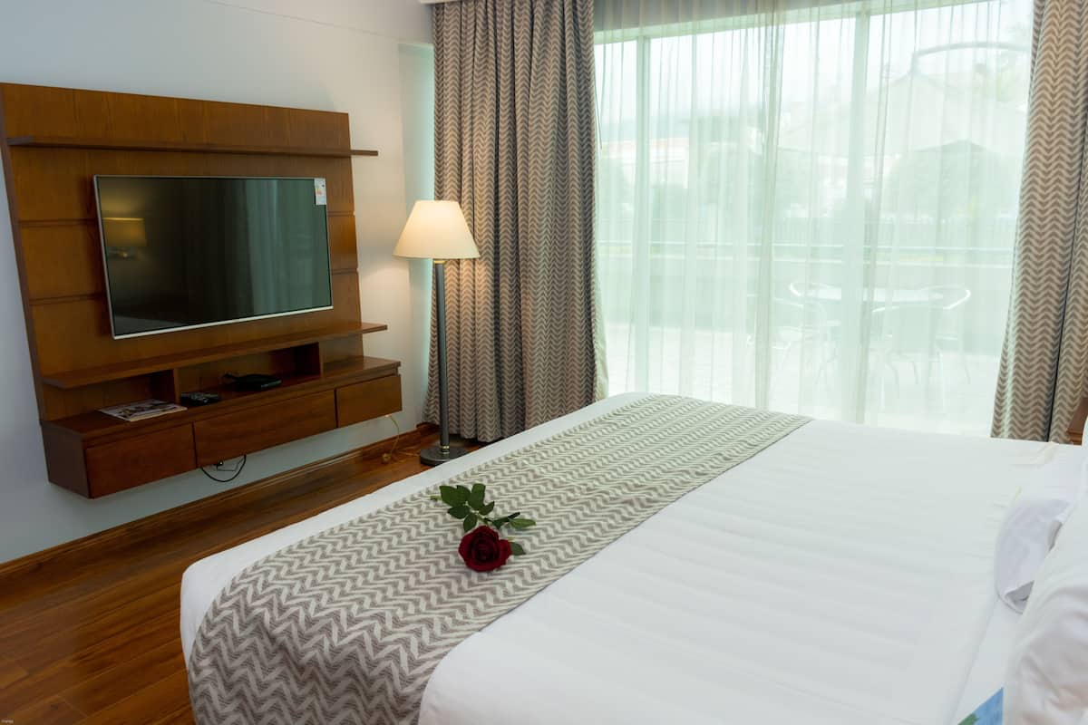 A hotel room with a large flat-screen TV, a bedside lamp, a bed with a patterned bedspread, and a window with curtains.