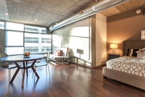 A modern hotel room with a large bed, a dining table, and a sofa. There is a window providing natural light and a view of the city. - Tripbz Mello Lofts (Los Angeles)