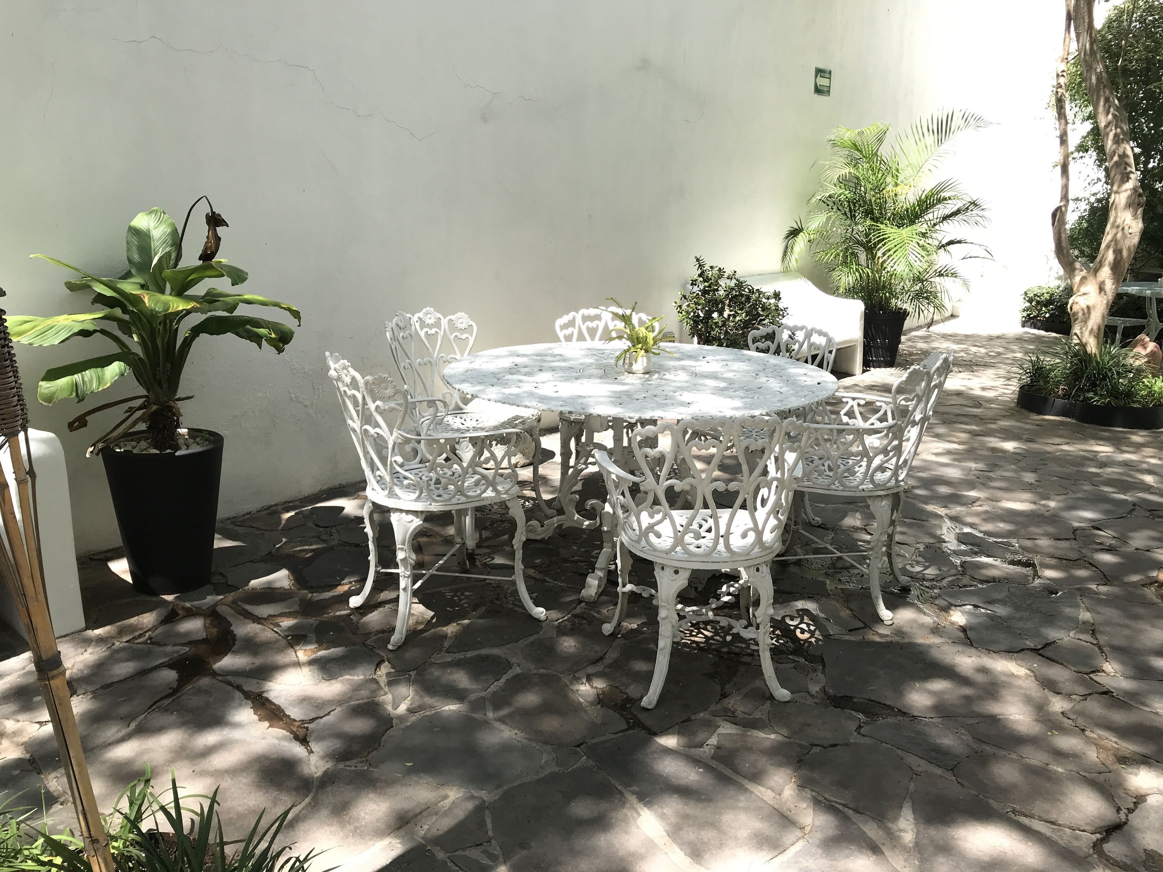A round outdoor table with white metal chairs, surrounded by potted plants and a stone-tiled floor.
