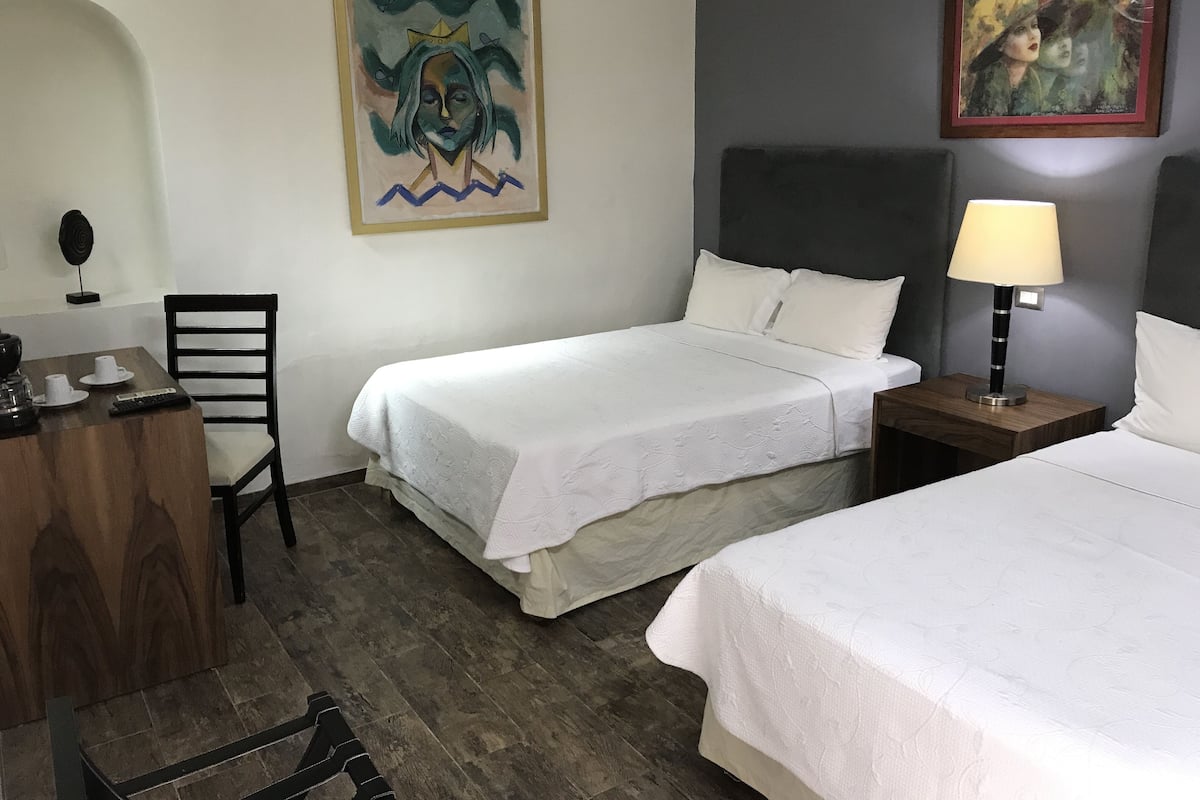 A hotel room with two single beds, a wooden desk with a chair, a lamp, and two paintings on the walls.