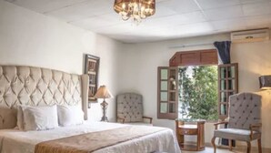 A bedroom with a tufted headboard, a bed with white linens, a bedside table with a lamp, a chair, a small wooden table, and a view of greenery through a window. - Casa Monraz (Guadalajara)