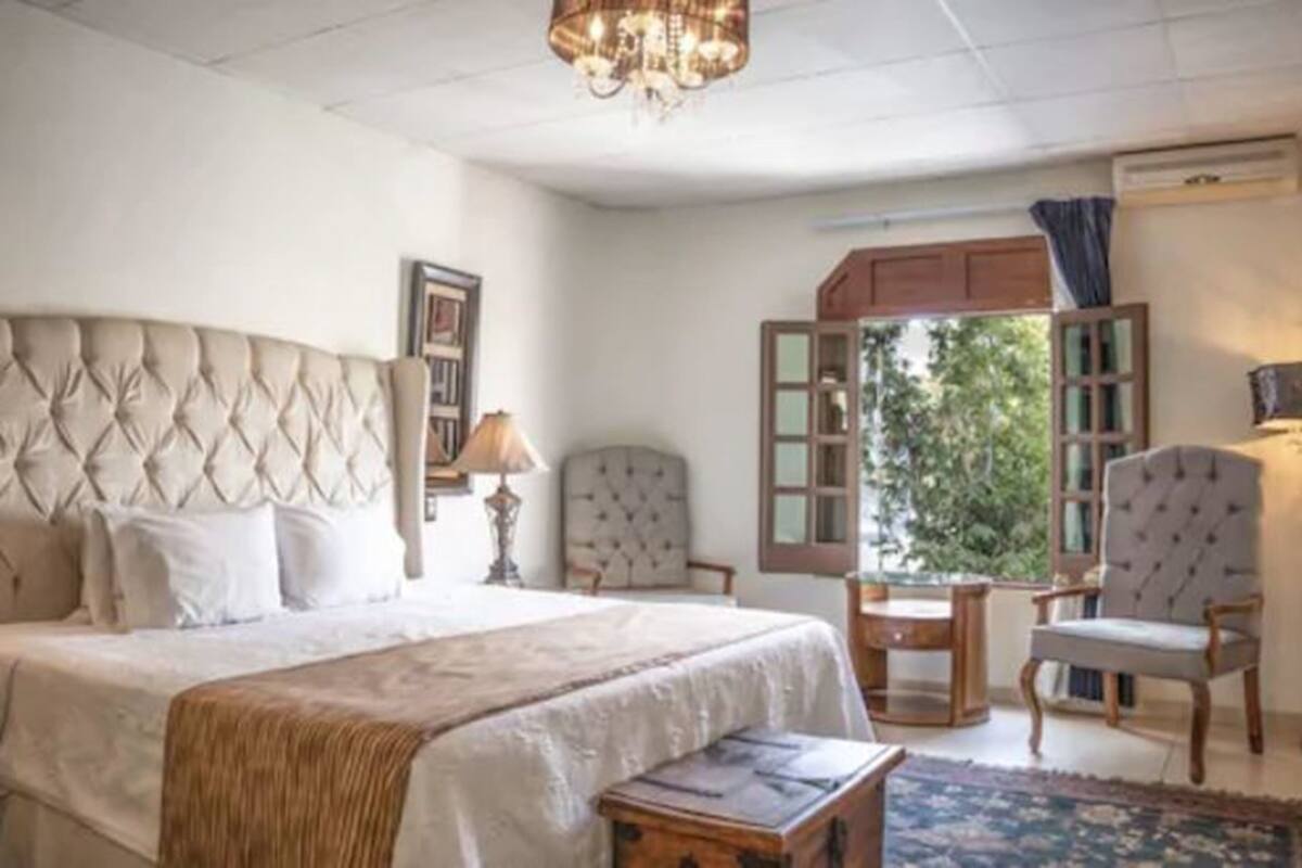 A bedroom with a tufted headboard, a bed with white linens, a bedside table with a lamp, a chair, a small wooden table, and a view of greenery through a window.