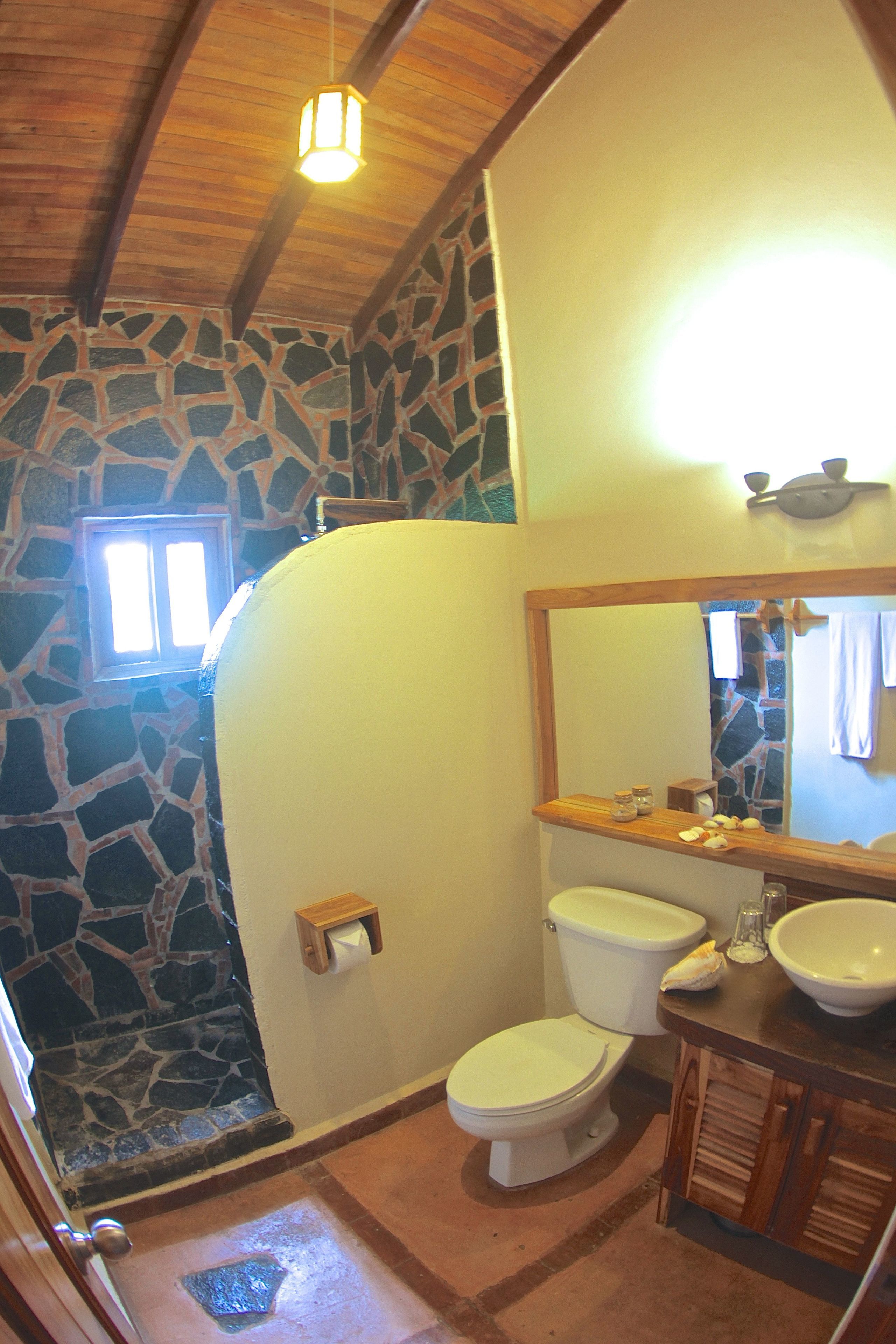 A bathroom with a stone wall, a wooden ceiling, a white toilet, a sink, and a mirror.
